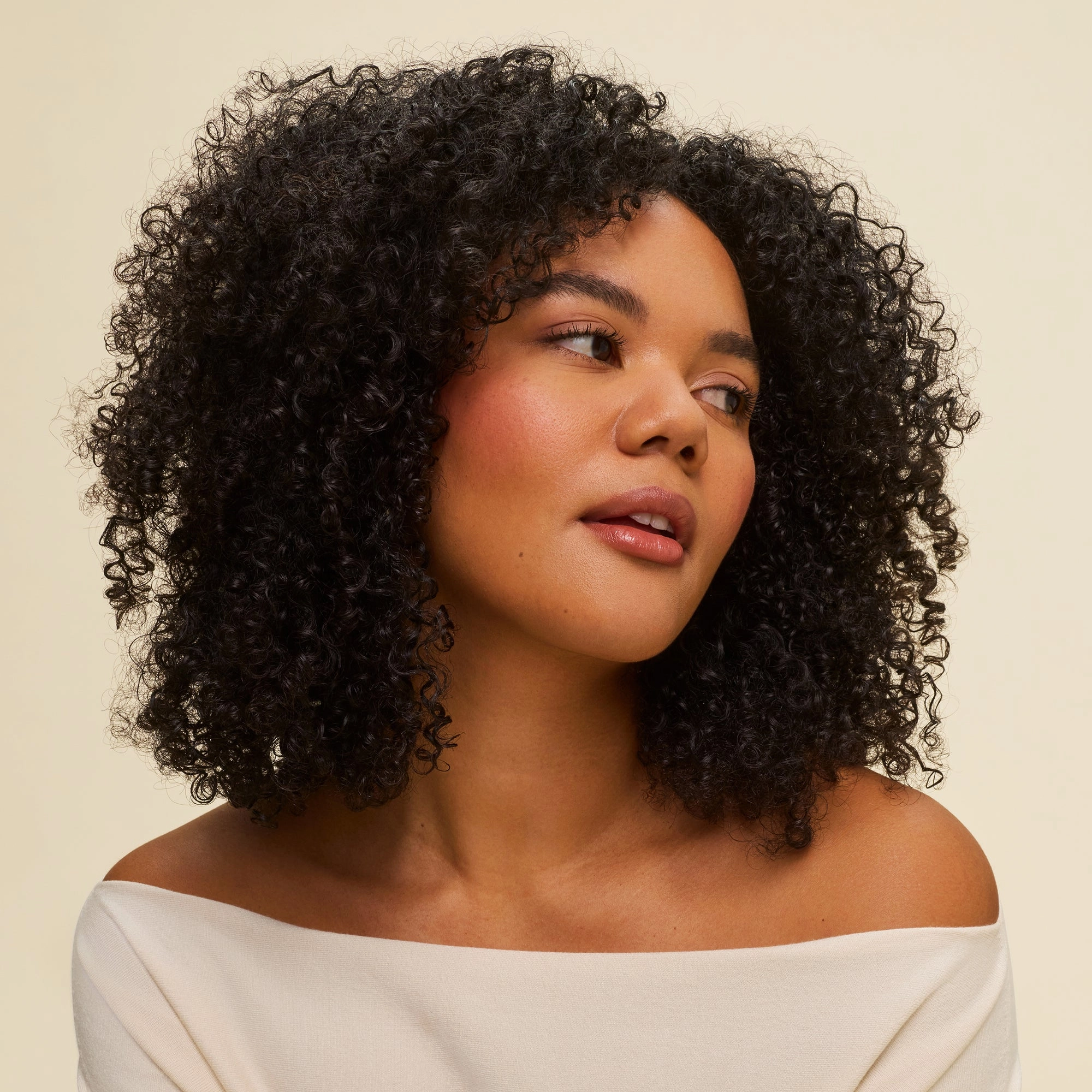 A woman with curly dark hair styled using Curl Defining Gel looks to the side against a plain light background