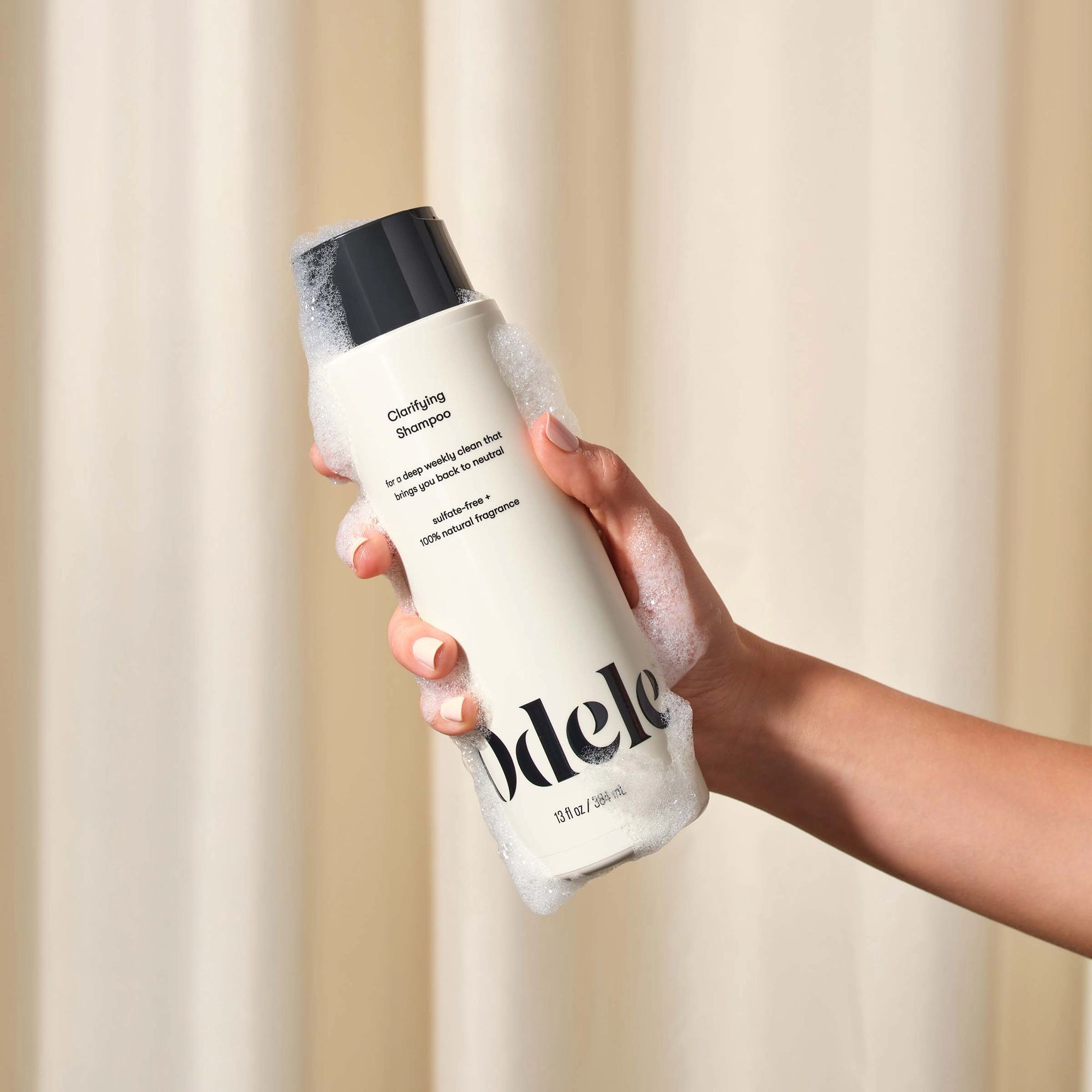 A hand covered in soap suds holds a white bottle of Odele Beauty Clarifying Shampoo with a black cap