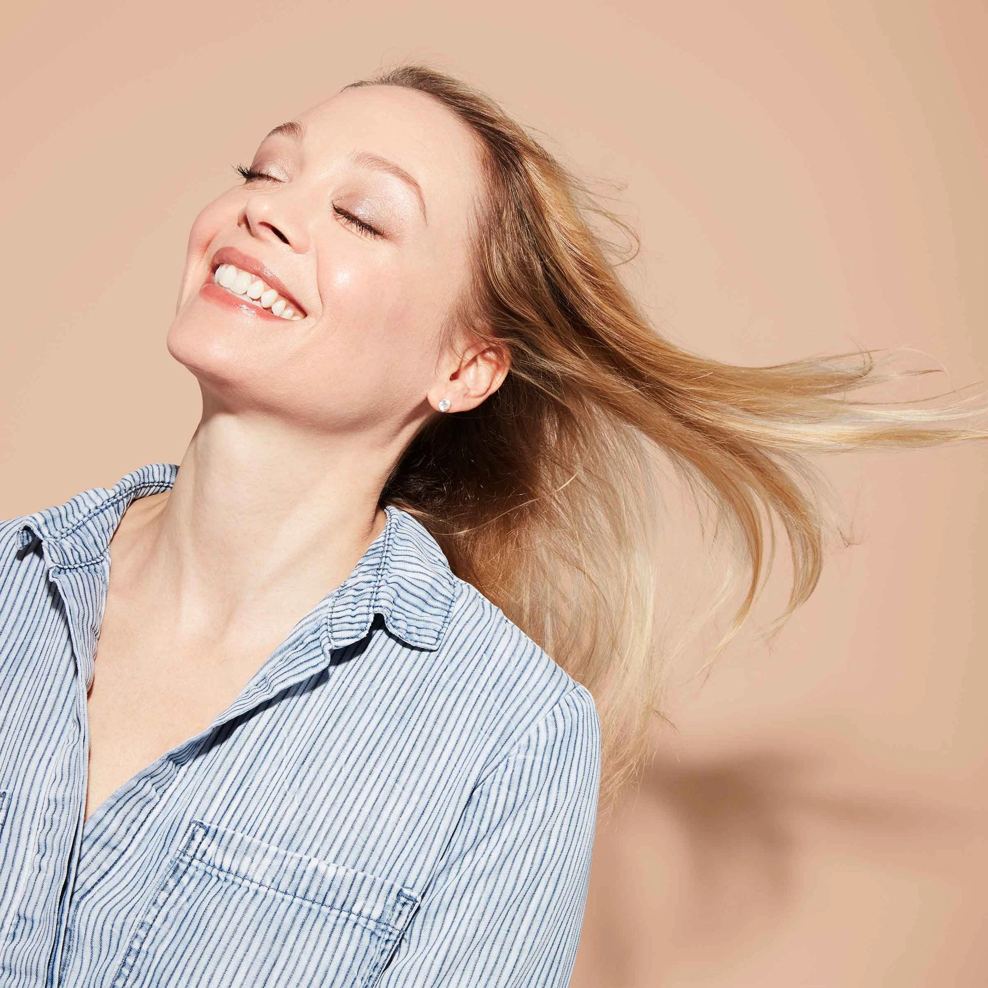 A smiling woman, eyes closed, tilts her head back and tosses her long, blonde hair over her shoulder