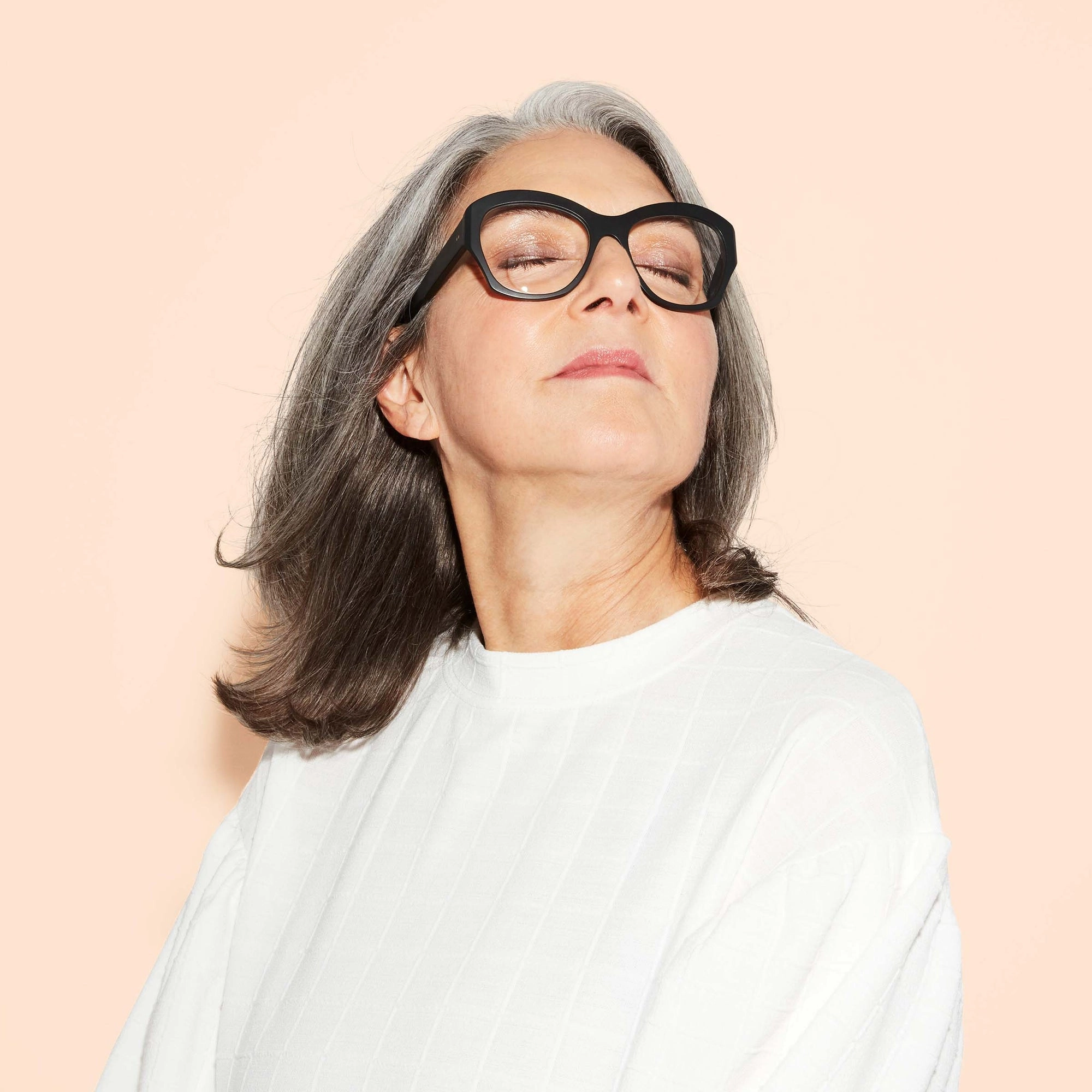 A confident older woman with gray hair and glasses, wearing a white top, poses with her head tipped upwards
