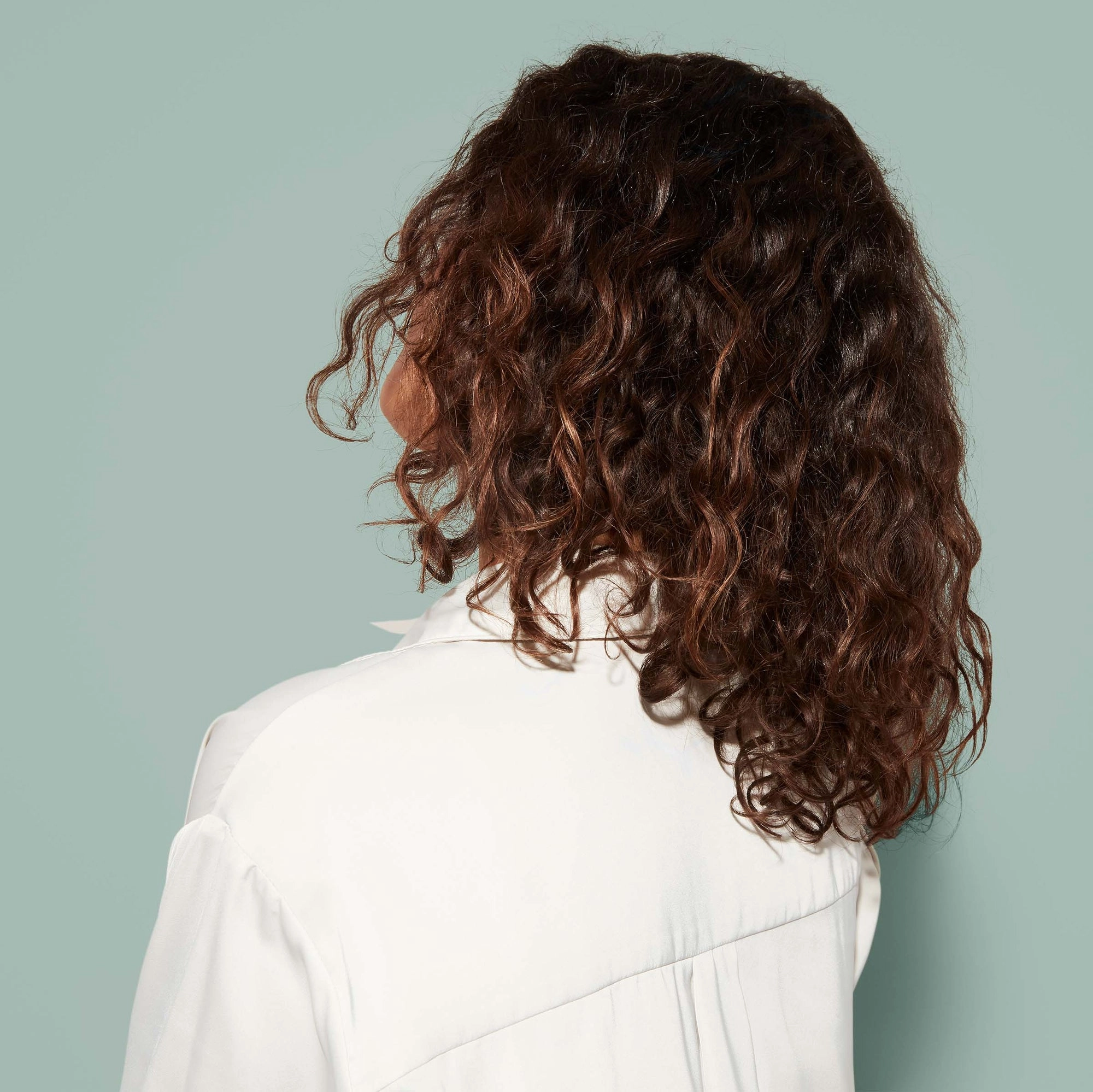 A young woman with shoulder-length brown waves wears a white shirt and is shown from behind