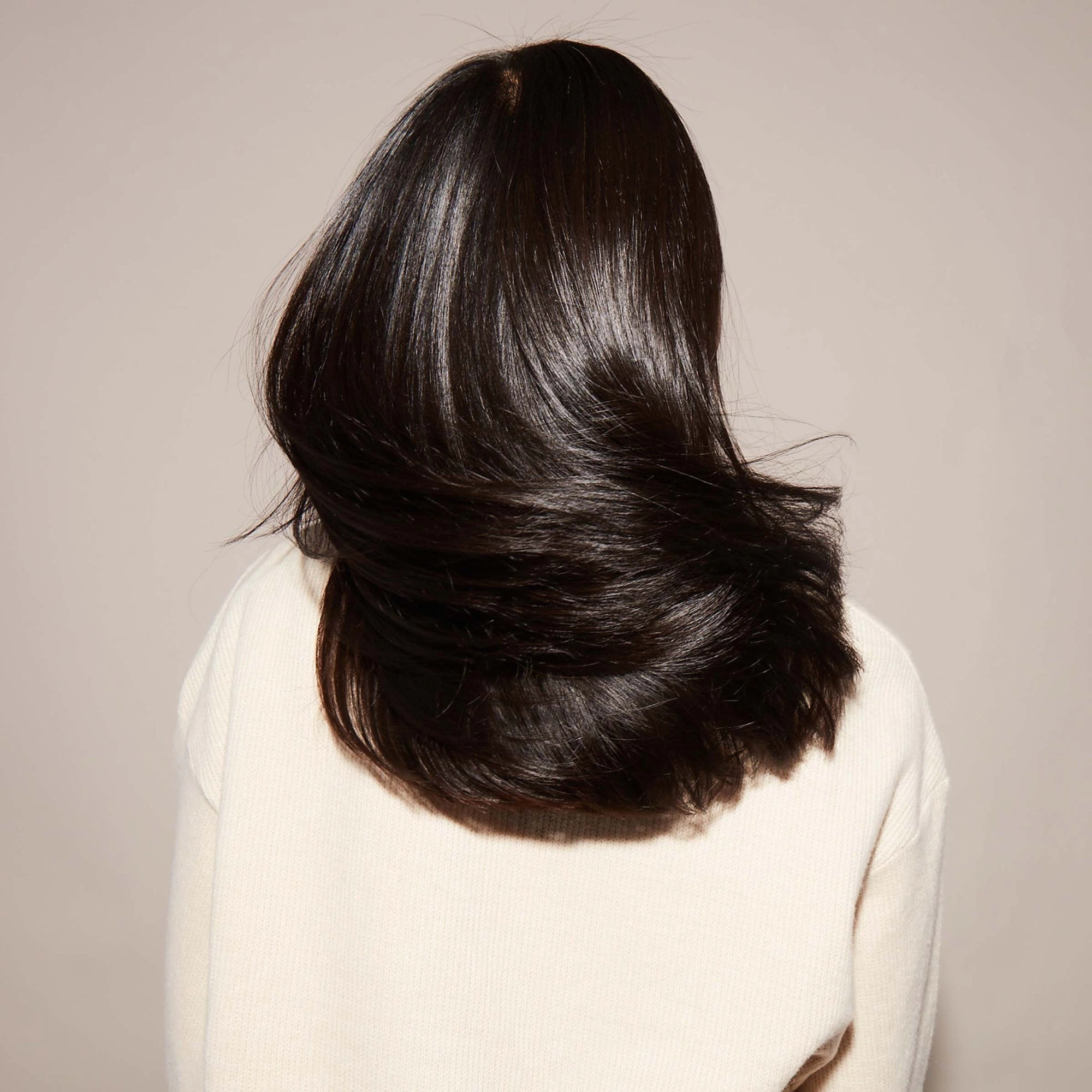 A woman with straight, shiny black hair faces away from the camera and swishes her hair back and forth